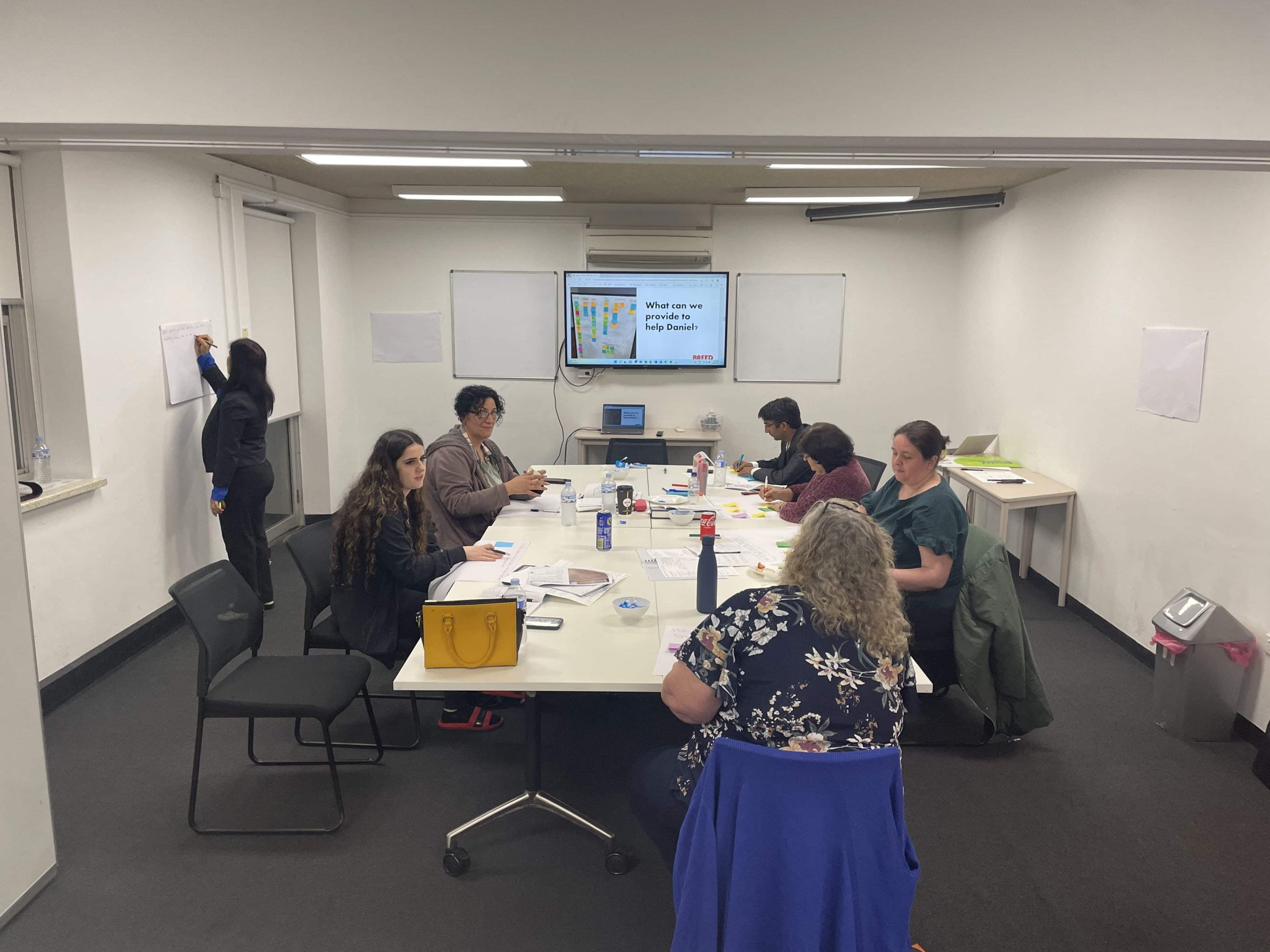 People are seated around a conference table in a meeting room, engaging in work. A presentation is displayed on a screen at the front of the room. One person is writing on a paper posted on the wall.