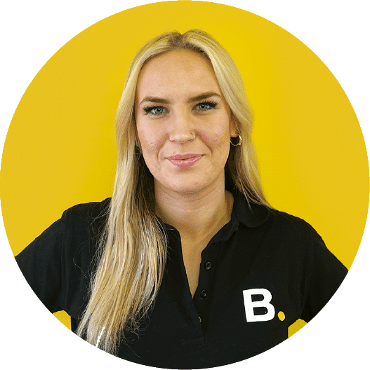 About 4 Person with long blonde hair wearing a black shirt with a white "B" logo stands against a yellow background.
