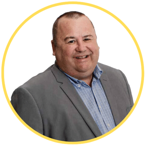 About 6 A man in a gray suit jacket and blue shirt smiling, enclosed in a circular yellow border.