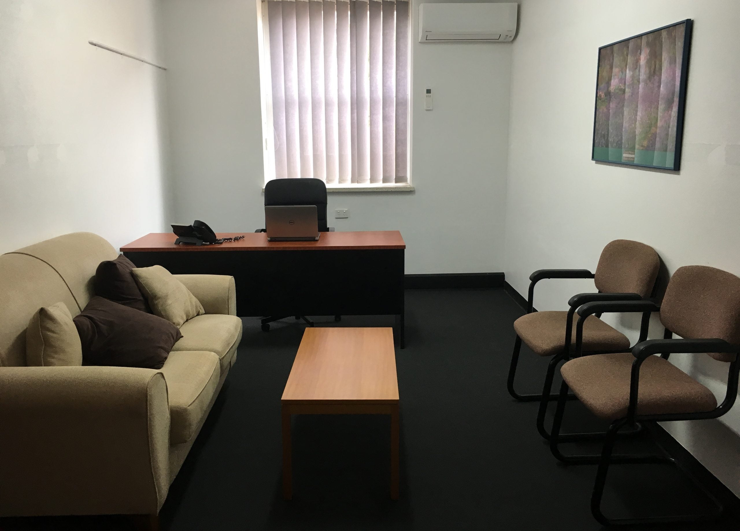 An office with a desk, chair, sofa, coffee table, and two additional chairs. The room has a window with vertical blinds, air conditioning, and a framed picture on the wall.