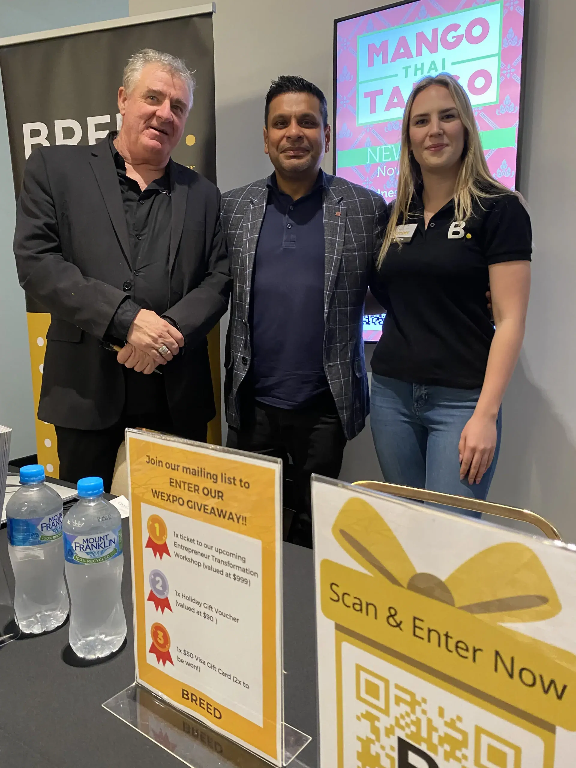 Three people are standing behind a table with promotional materials for BREED. The table displays water bottles, a sign about a giveaway, and a QR code for entry. A screen in the background reads "Mango Thai Tapas".