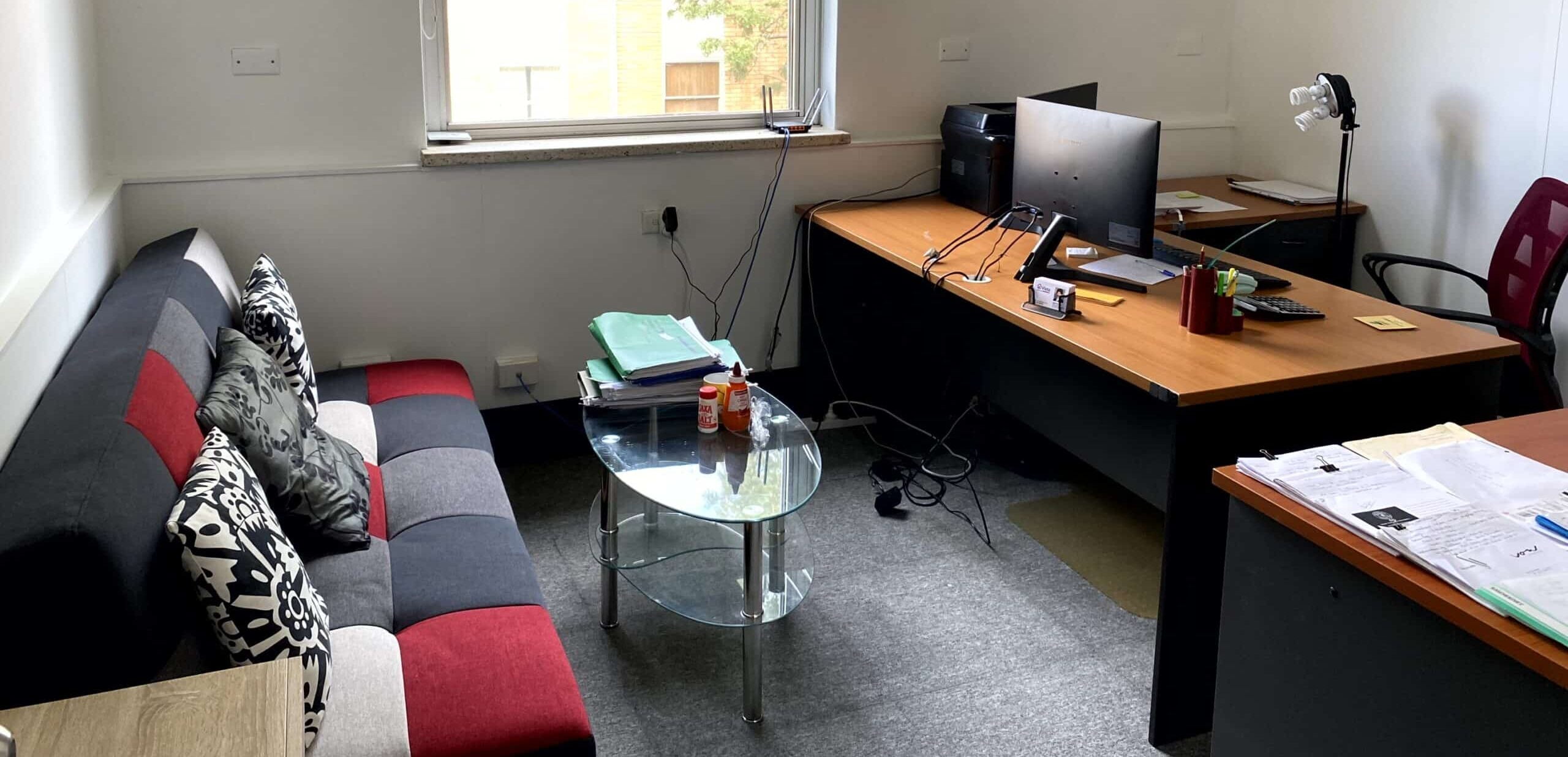 A small office with a desk, computer, chair, and various office supplies. There is a sofa, glass coffee table, and a large window letting in natural light. Wall decor includes framed artwork.