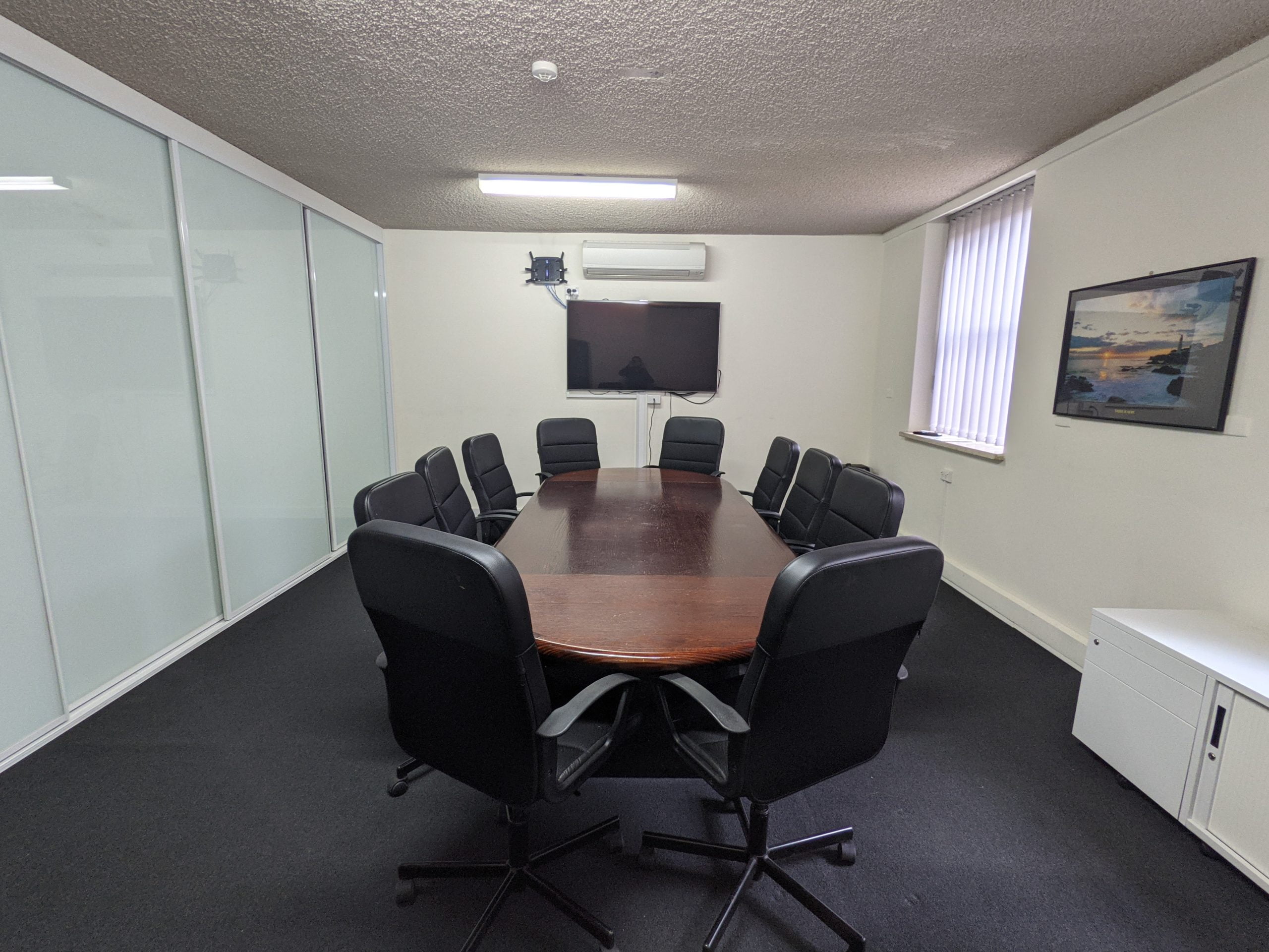 A conference room with a long oval table surrounded by ten black office chairs. There is a wall-mounted TV, a window with blinds, a white cabinet, and a painting on the wall.