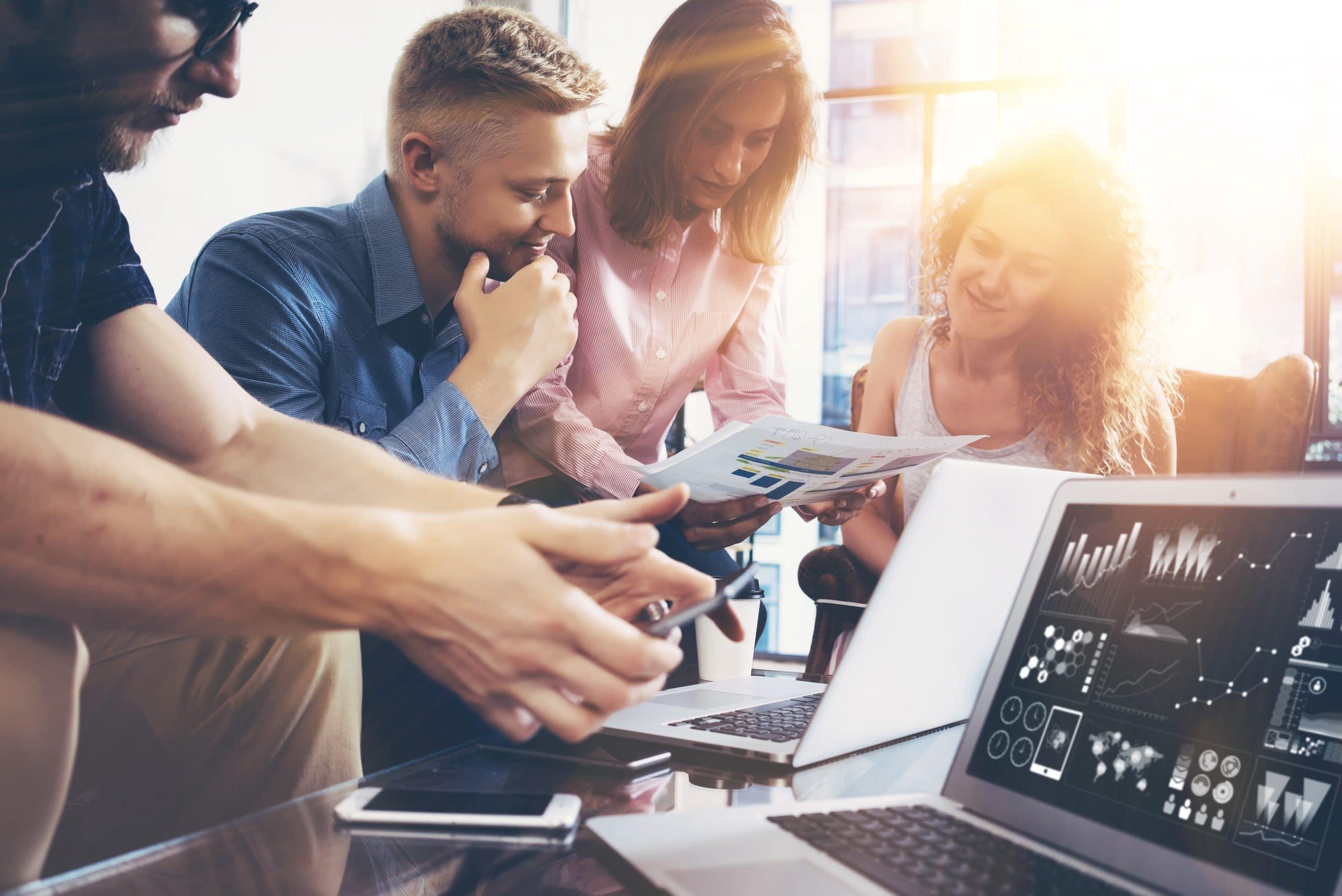 A group of four people gathers around a table, engaging in discussion while examining documents and using laptops. Sunlight streams through the windows in the background.