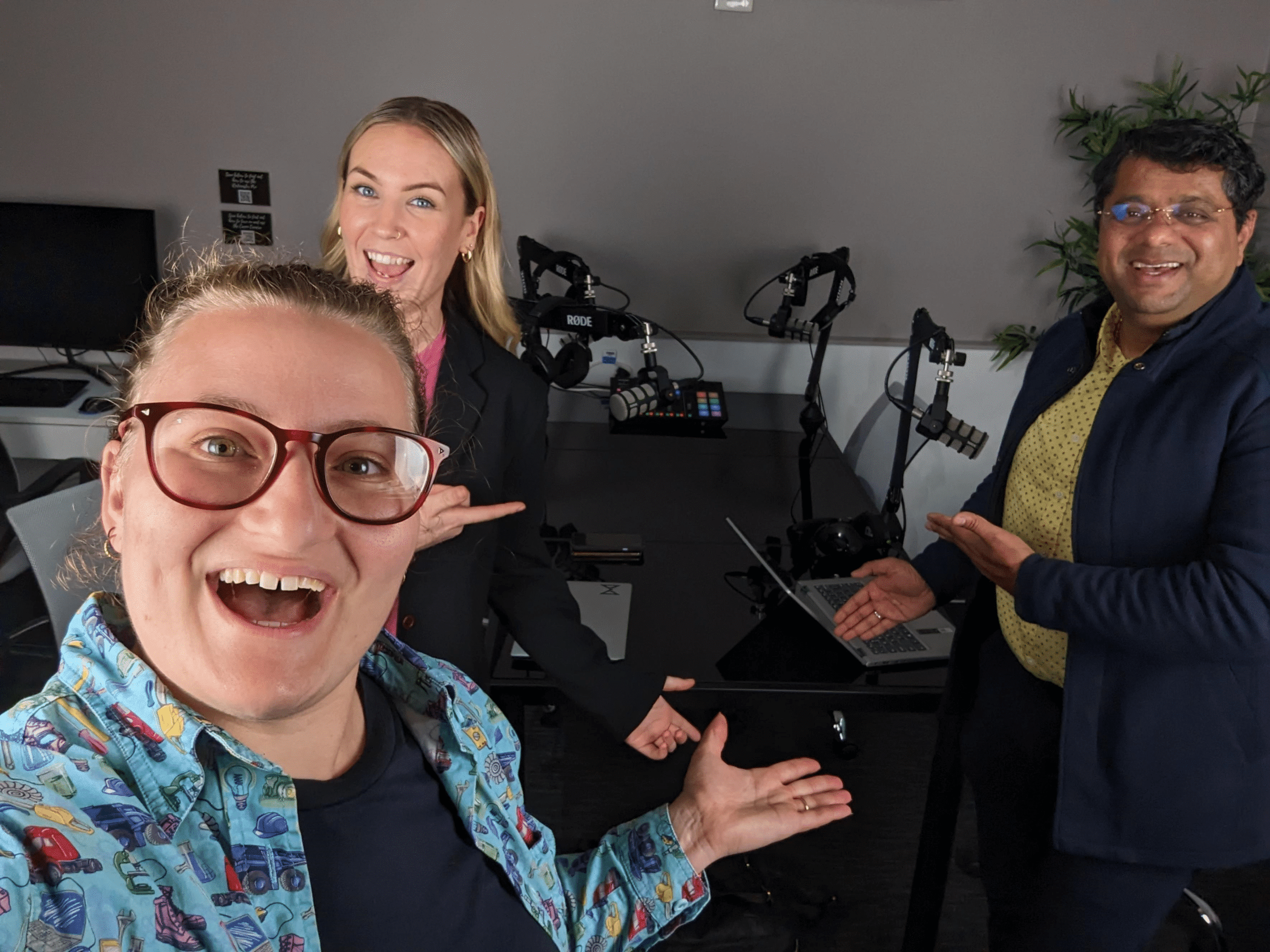 Three people in a recording studio, smiling and gesturing towards the podcast equipment on a table.