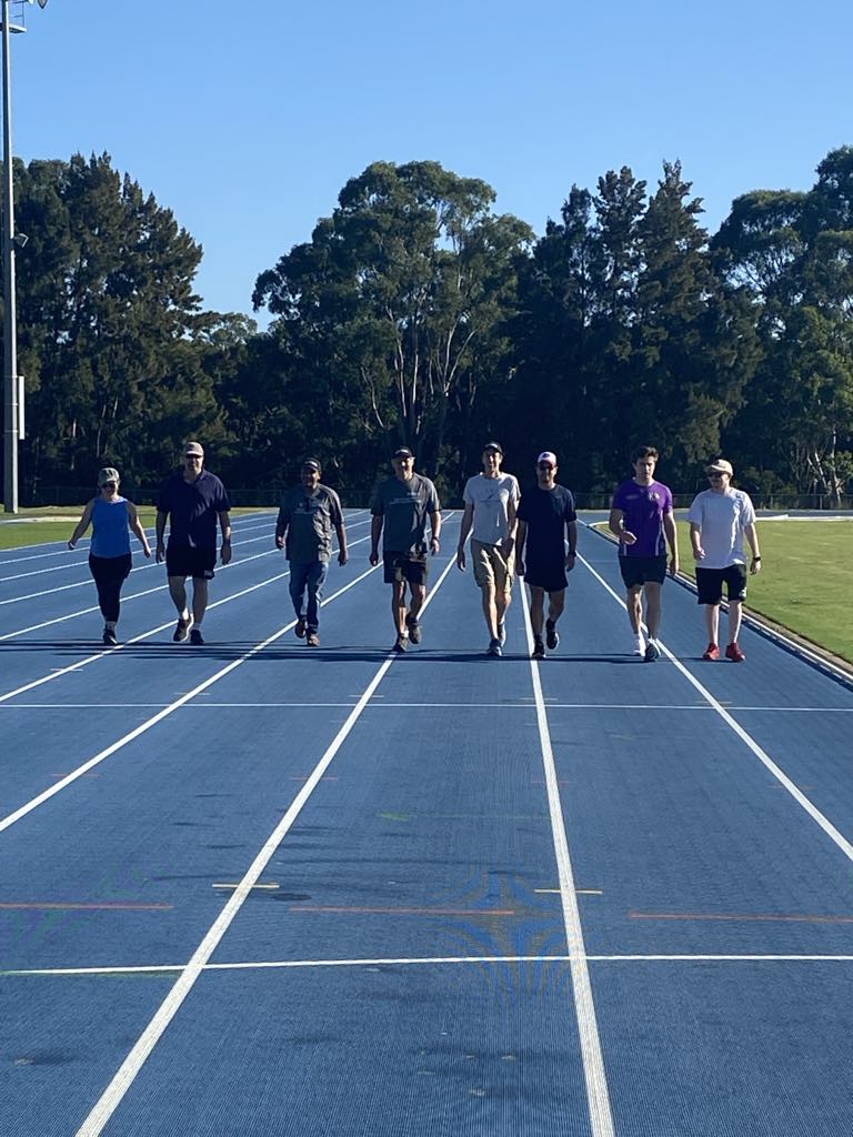 Seven people are walking side by side on a blue running track surrounded by trees and clear sky, enjoying the fresh air as part of their charity walk registration event.