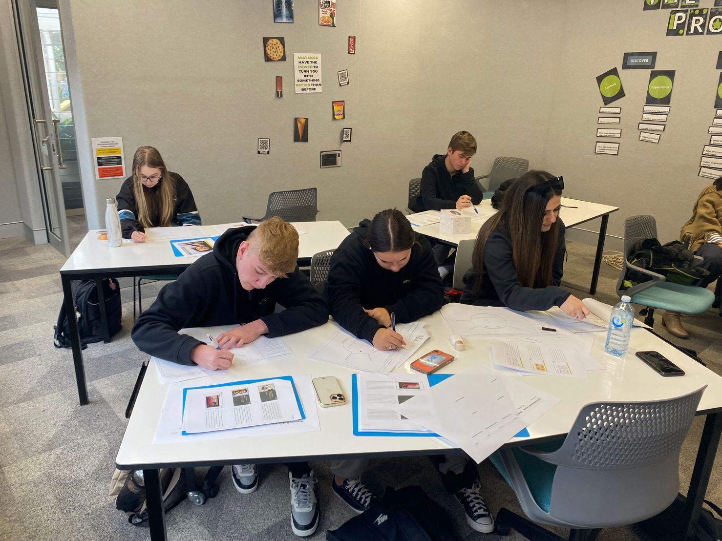 Students sit at desks in a classroom, focusing on writing and drawing assignments. Various educational posters are visible on the walls.