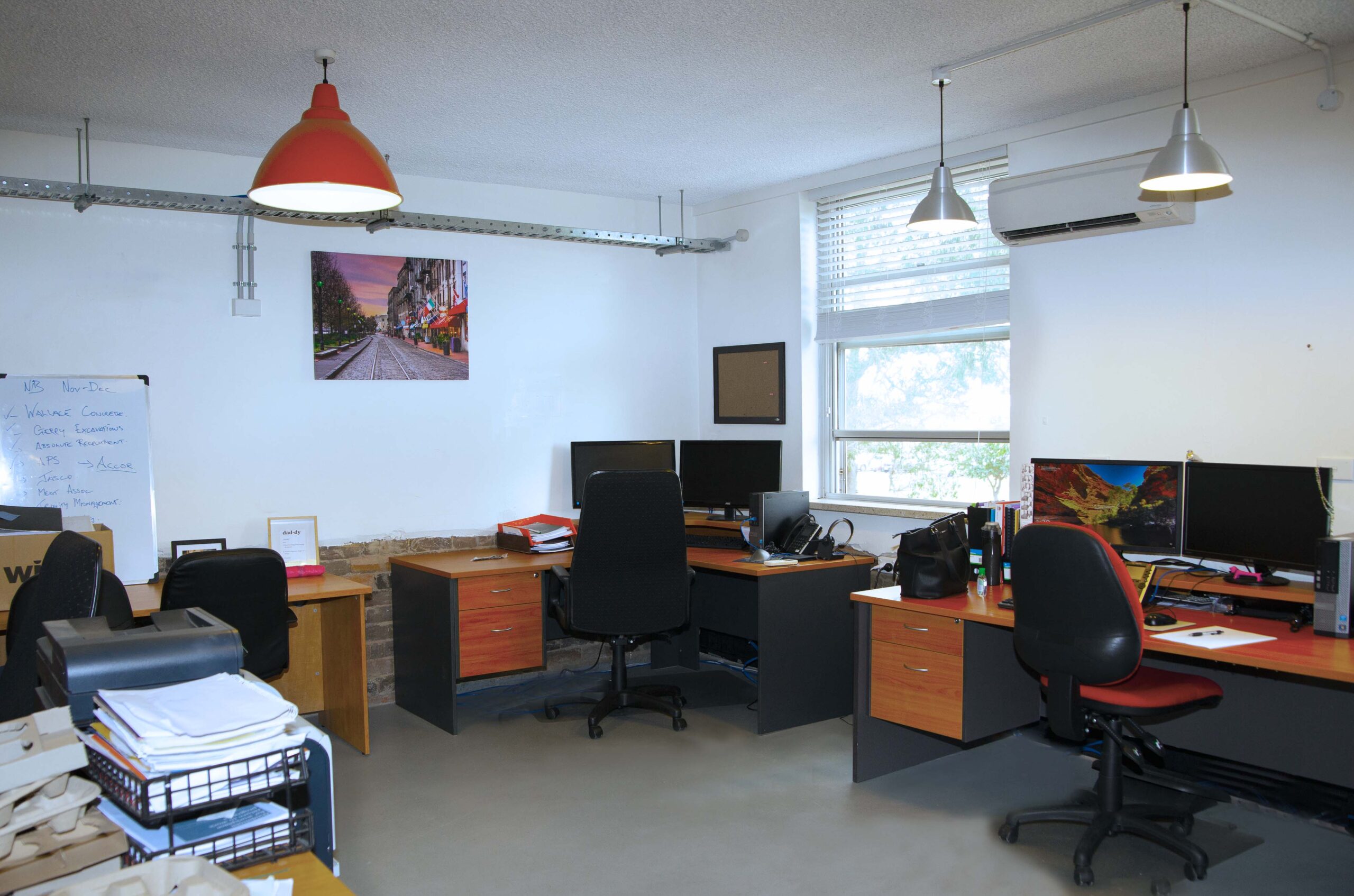 A small office space with multiple desks equipped with computers, chairs, and office supplies. There are wall-mounted lights, a window, and a whiteboard with notes. A framed photo hangs on the wall.