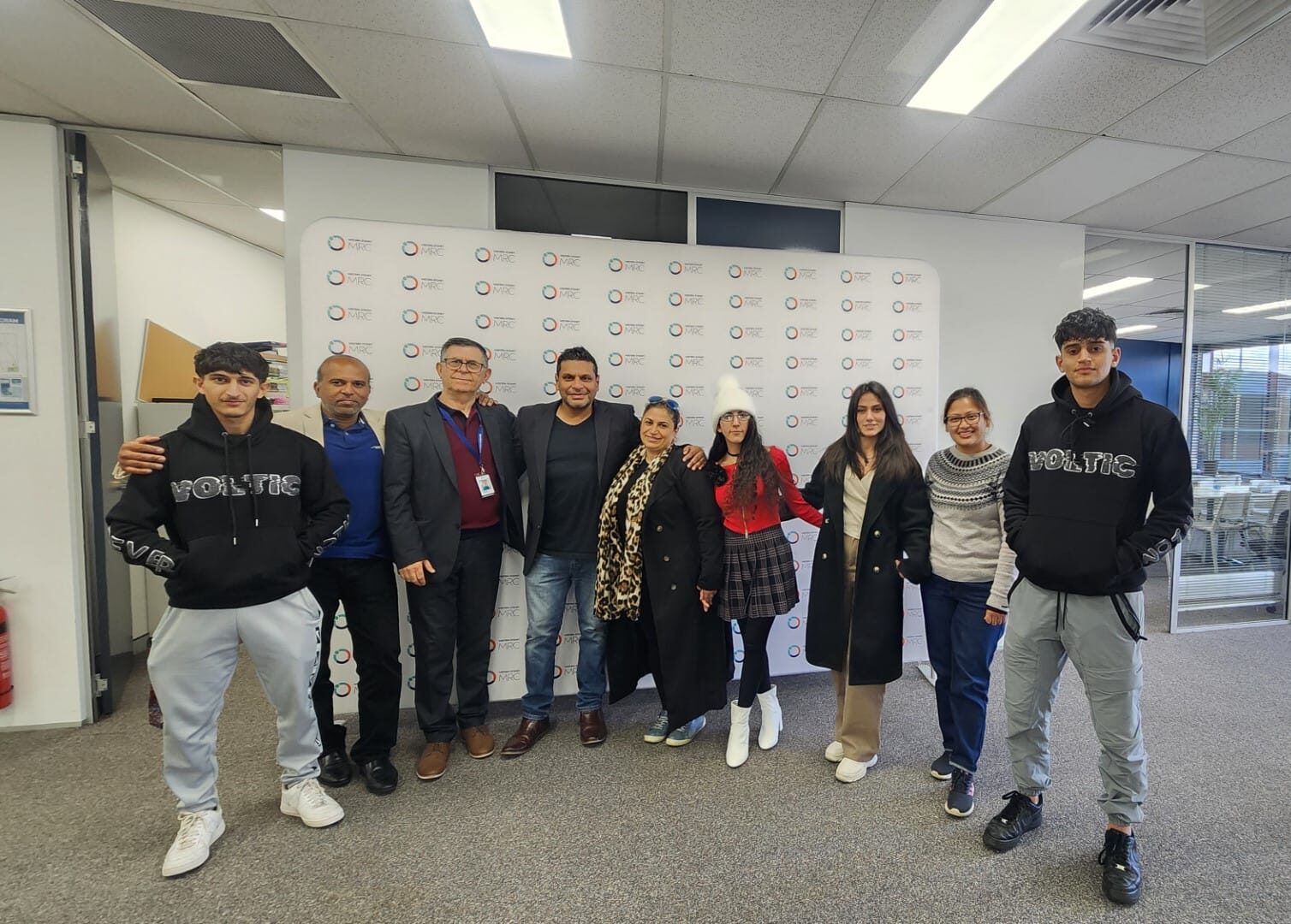A group of eight people standing in an office room with a branded backdrop, facing the camera.