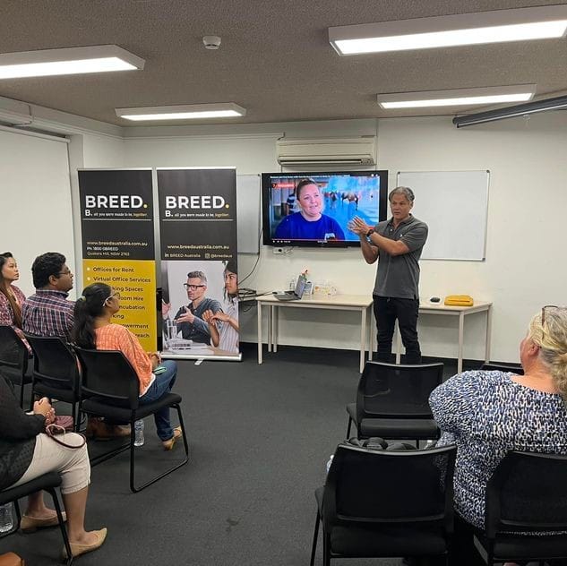 A person is giving a presentation in a classroom setting with a screen showing another person. Attendees are seated and two banners labeled "BREED" are displayed in the background.