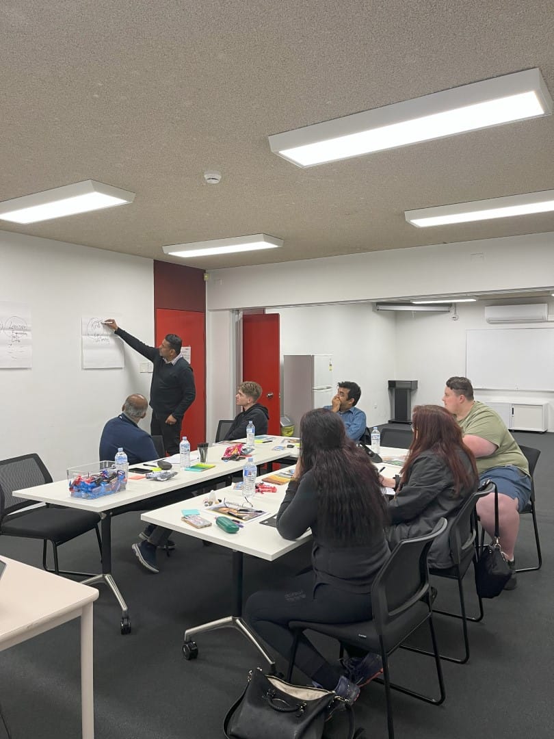 A group of people seated at tables watches a person stand and point to charts on a wall in a meeting room. Various items, such as snacks and drinks, are on the tables.
