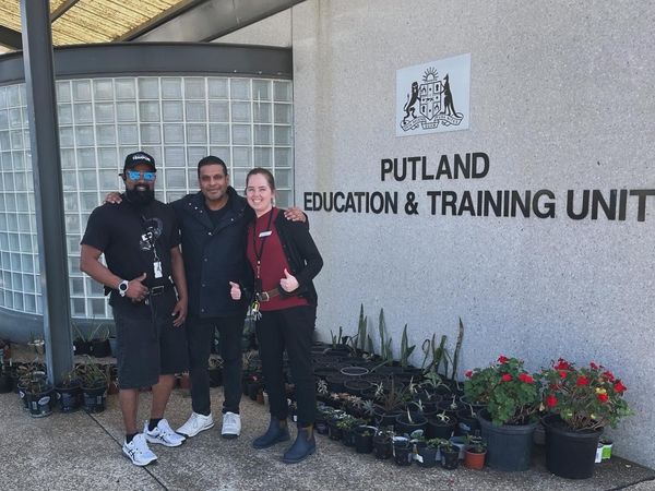 Three people stand smiling in front of the Putland Education & Training Unit building, surrounded by potted plants.