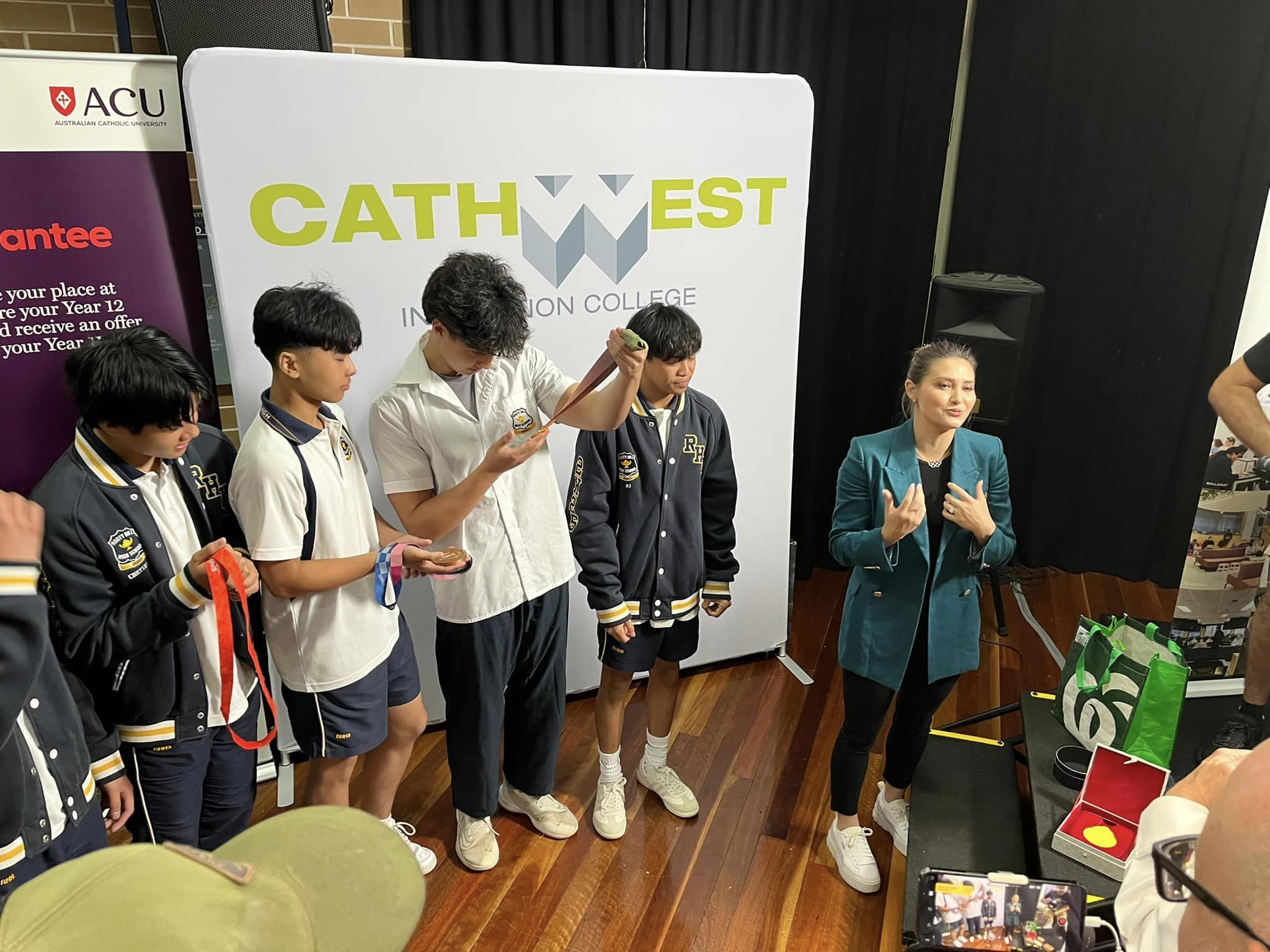 A group of students in uniforms stand near a woman gesturing with her hands in front of a CathWest Innovation College banner.