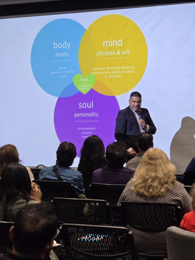 A presenter stands in front of a small business audience with a projected slide showing overlapping circles labeled "body," "mind," and "soul.