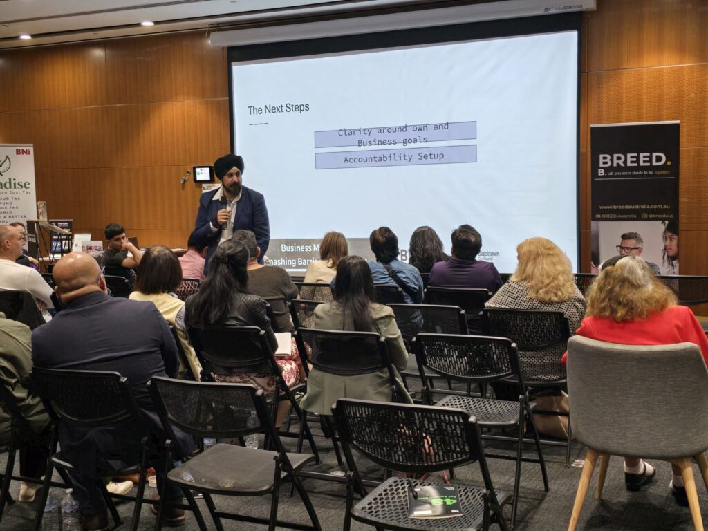 A man presents a slide titled "The Next Steps" to an audience in a conference room, highlighting strategies essential for small business growth.