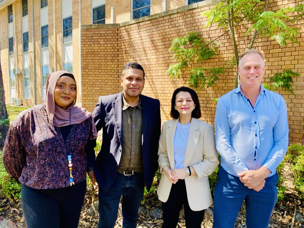 Four people standing outdoors in front of a brick building, three are wearing business attire and one is in a casual shirt. There is greenery in the background.