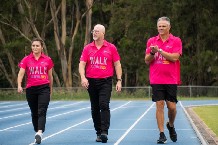 Three people in pink shirts stroll along a blue running track, surrounded by trees, at the vibrant Walk Festival in Western Sydney.