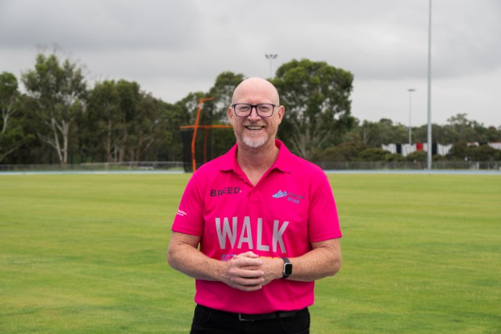 A man wearing glasses and a pink "WALK" shirt smiles while standing on a sports field in Western Sydney, with trees and a cloudy sky in the background, promoting youth mental health awareness.