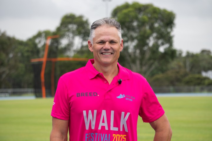 A person wearing a pink "Walk Festival 2025" shirt stands smiling on a grassy field in Western Sydney, with trees providing a serene backdrop.