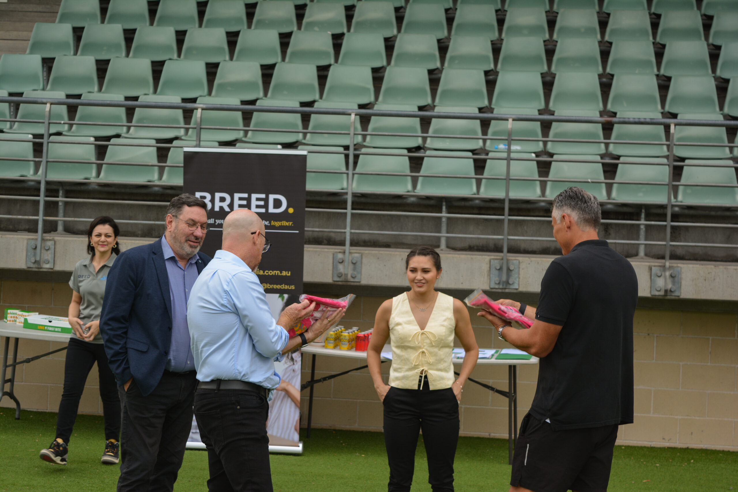 A group of people gather on a sports field in Western Sydney, talking and smiling. Some hold pink paper bags, likely from the walk festival promoting youth mental health awareness. Bleachers and a "BREED" banner are visible in the background, capturing the lively community spirit.