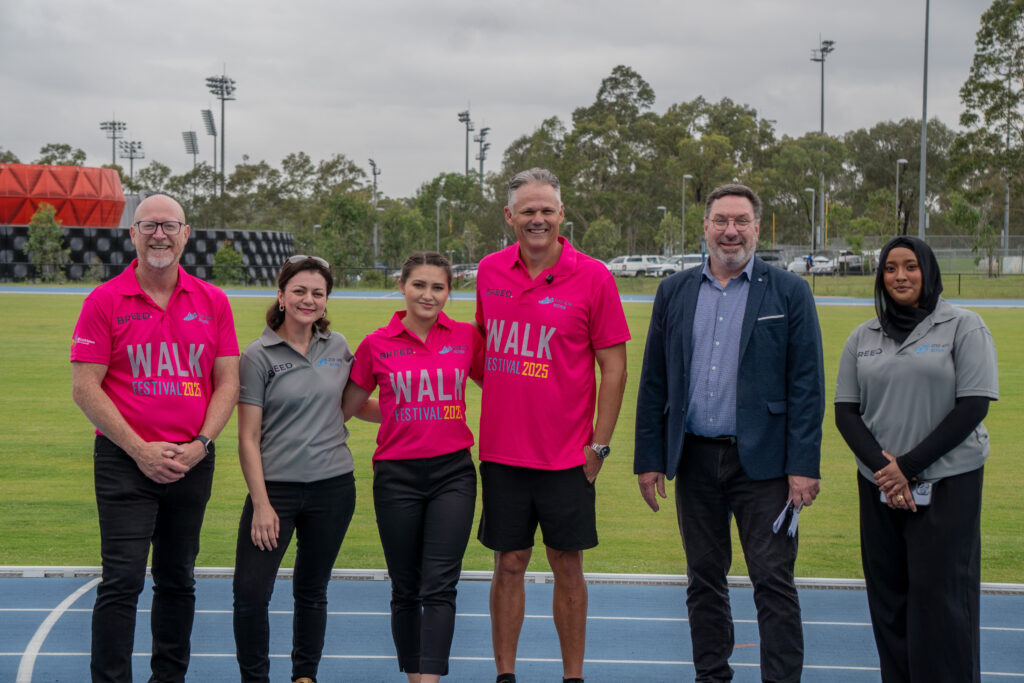 A group of six people stand on an outdoor track in Western Sydney. Two wear pink "WALK Festival 2023" shirts. Trees and sports facilities can be seen in the background, creating a vibrant setting that highlights their support for youth mental health initiatives.