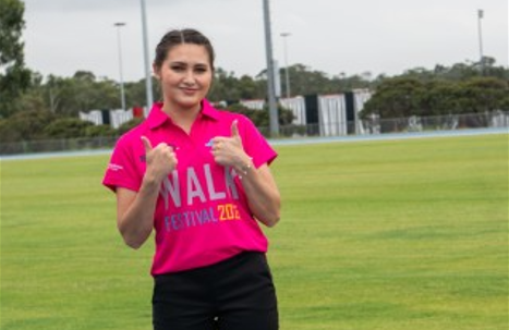 A person in a pink "Walk Festival 2023" shirt gives two thumbs up while standing on a grassy field. In the background, trees and poles hint at the lively spirit of Western Sydney, underscoring an event devoted to promoting youth mental health.