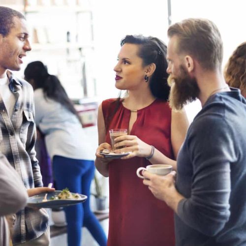 A group of people are in conversation at a social gathering. They are holding drinks and plates of food. The setting appears to be indoors with large windows in the background.