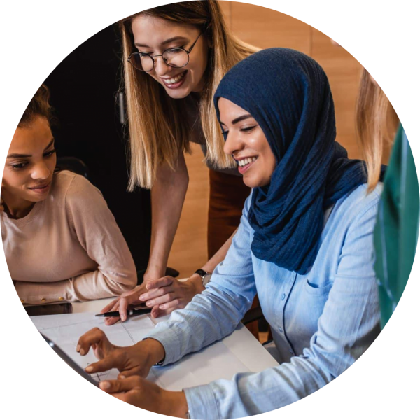 Three women are gathered around a table, smiling and looking at documents or a device. One woman wears glasses and another wears a hijab. They appear to be collaborating in an office setting.