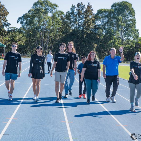 A group of people walking on a blue running track, surrounded by trees and tents, some wearing black shirts and one person waving.