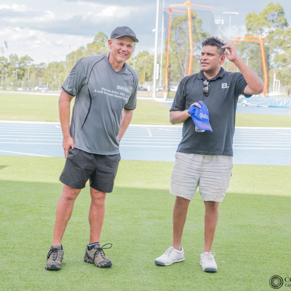 Two men stand on a sports field, one holding a blue cap. They are dressed in sports attire and engaged in conversation.