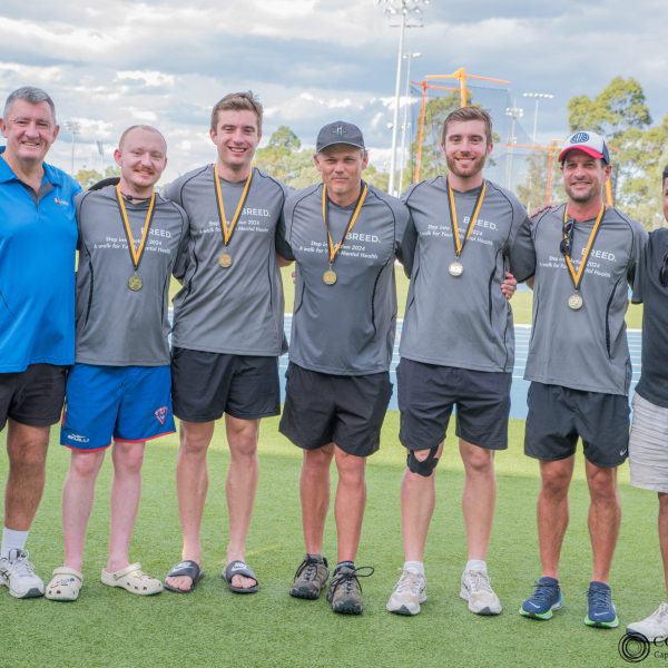 A group of seven men standing together on a track field, five wearing matching gray shirts with medals around their necks, flanked by two others in casual attire.
